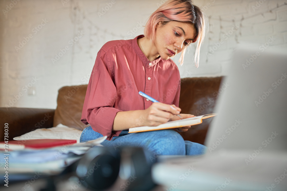 Concentrated young woman copywriter with pink hair working from home making notes in copybook. Cute student girl putting down ideas, writing essay, having focused facial expression, sitting on couch