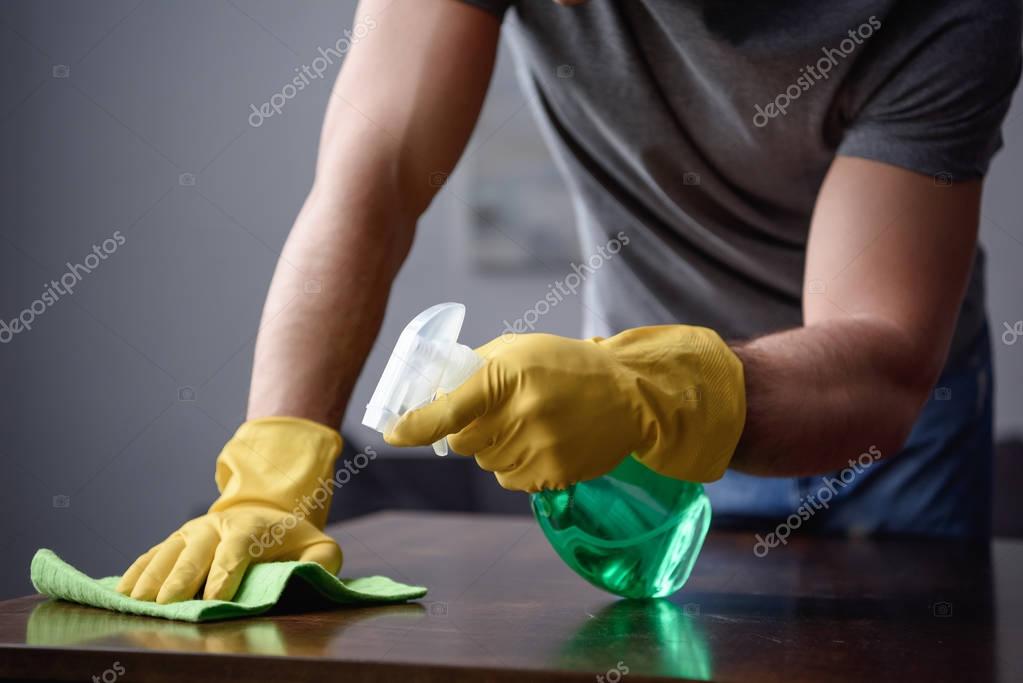 cropped image of man cleaning table in living room with spray bottle and rag