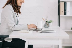 cropped view of smiling secretary using laptop while sitting at workplace in office