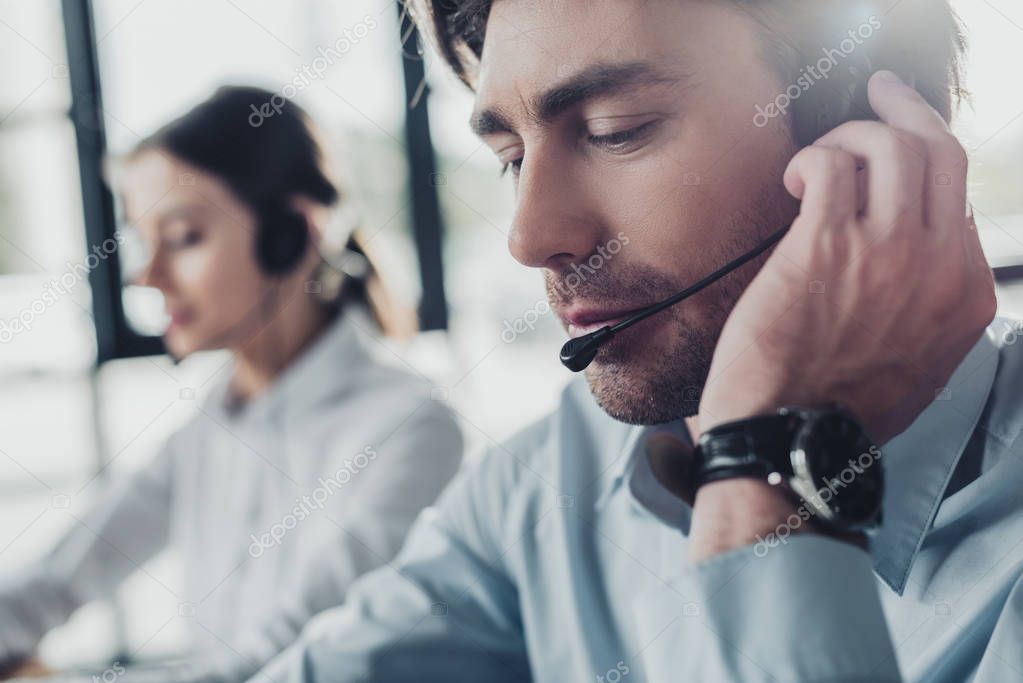 handsome call center manager in headphones with mike sitting at workplace while his colleague sitting on background