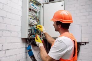 Handsome electrician in hardhat and gloves holding multimeter near electrical distribution box