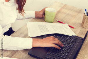 Mujer  trabajando con un ordenador escribiendo y con papeles y una taza de café sobre un escritorio de madera. Vista superior