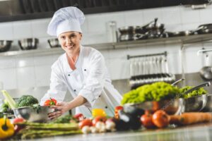 smiling attractive chef taking bowl with vegetables and looking at camera at restaurant kitchen