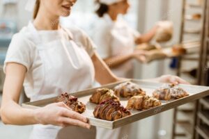 cropped shot of young baker holding tray with fresh croissants at baking manufacture