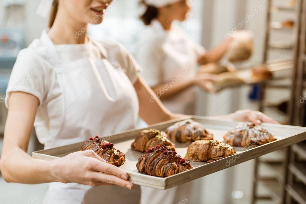 cropped shot of young baker holding tray with fresh croissants at baking manufacture