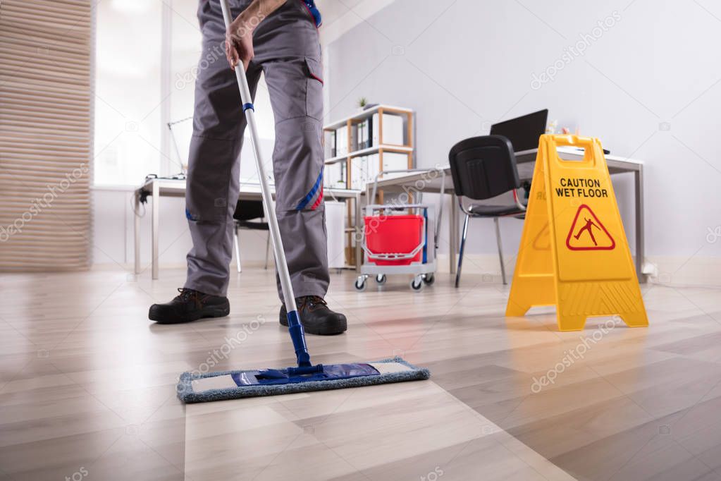 Low Section Of Male Janitor Cleaning Floor With Caution Wet Floor Sign In Office