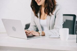 partial view of smiling secretary using laptop while sitting at workplace in office