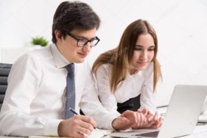 Young smiling businessman looking at laptop screen and making notes, businesswoman next to him. Concept of help.