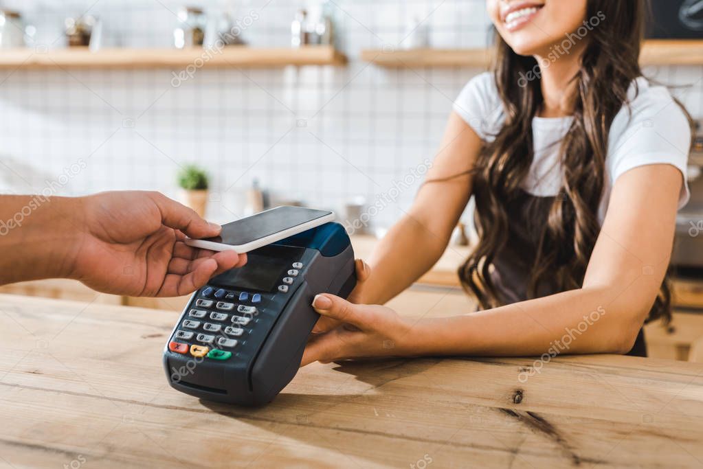 cropped view of cashier standing near bar counter in brown apron and holding terminal wile man paying with smartphone in coffee house