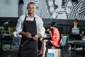 handsome young waiter in apron standing at cafe