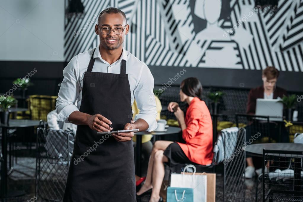 handsome young waiter in apron standing at cafe