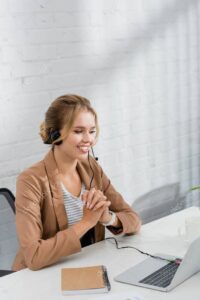 Cheerful female consultant in headset, looking at laptop while sitting at workplace