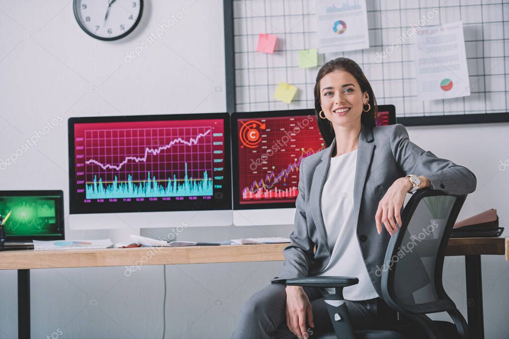 Smiling computer systems analyst looking at camera near charts on computer monitors on table