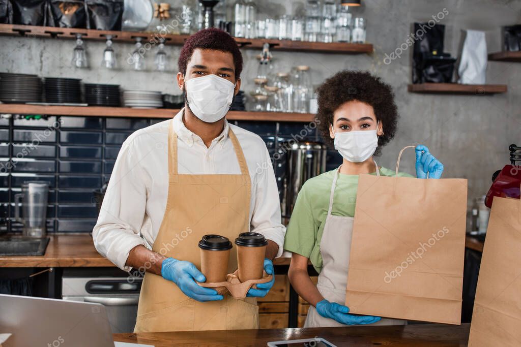 African american baristas in medical masks holding paper bag and coffee to go in cafe