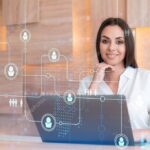 Attractive businesswoman in white shirt at workplace working with laptop to hire new employees for international business consulting. HR, social media hologram icons over office background