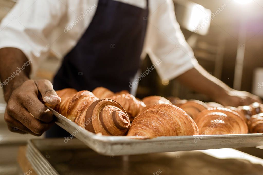 cropped shot of baker holding tray with croissants on baking manufacture