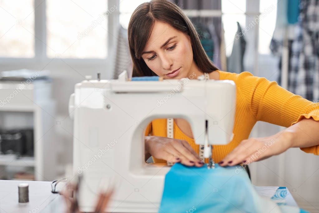 Focused charming caucasian fashion designer sitting in her studio and sewing beautiful evening dress.