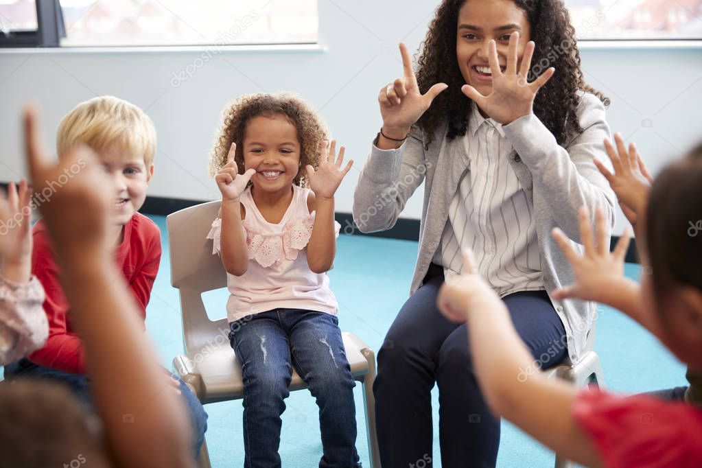 Front view of infant school children sitting on chairs in a circle in classroom, holding up their hands and learning to count with their female teacher