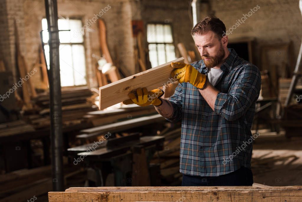 bearded carpenter in protective gloves checking wooden plank at sawmill