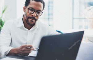 Dark skinned coworker wearing glasses and using laptop in modern office.African american man in white shirt working on workplace.Horizontal,blurred background